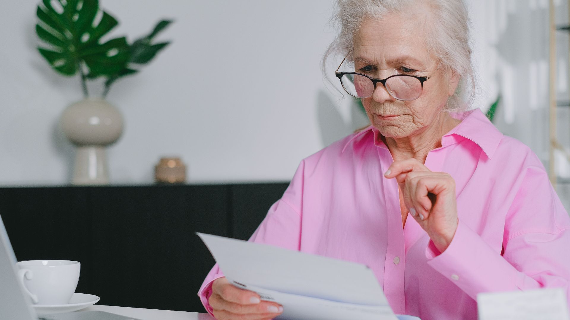 Elderly woman in pink shirt reading documents at home workspace.
