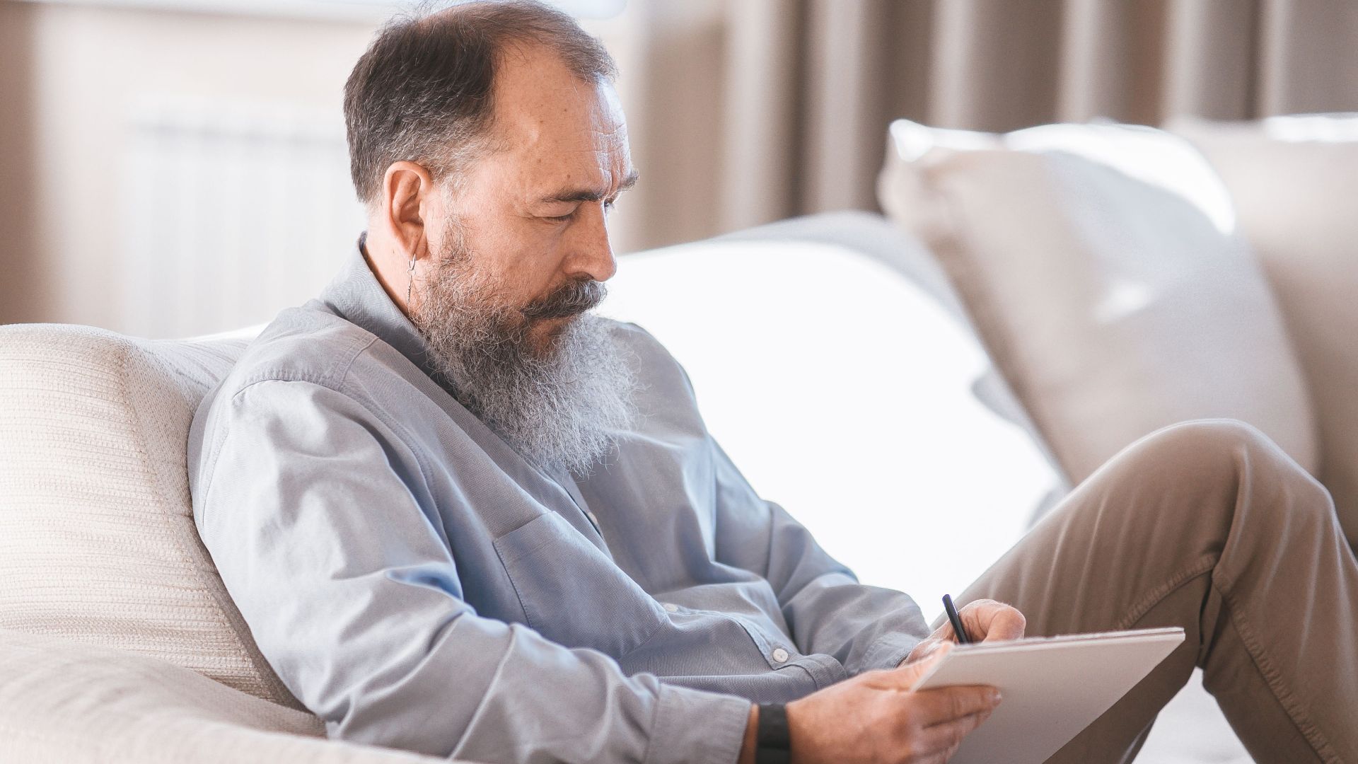 Elderly man with a beard sitting on a sofa, reading on a tablet indoors.