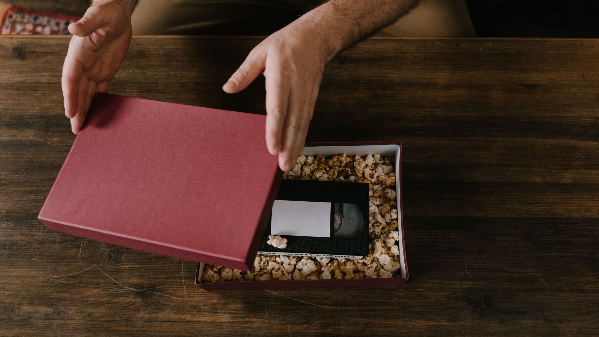 Hands opening a maroon gift box containing a VHS tape cushioned in popcorn.