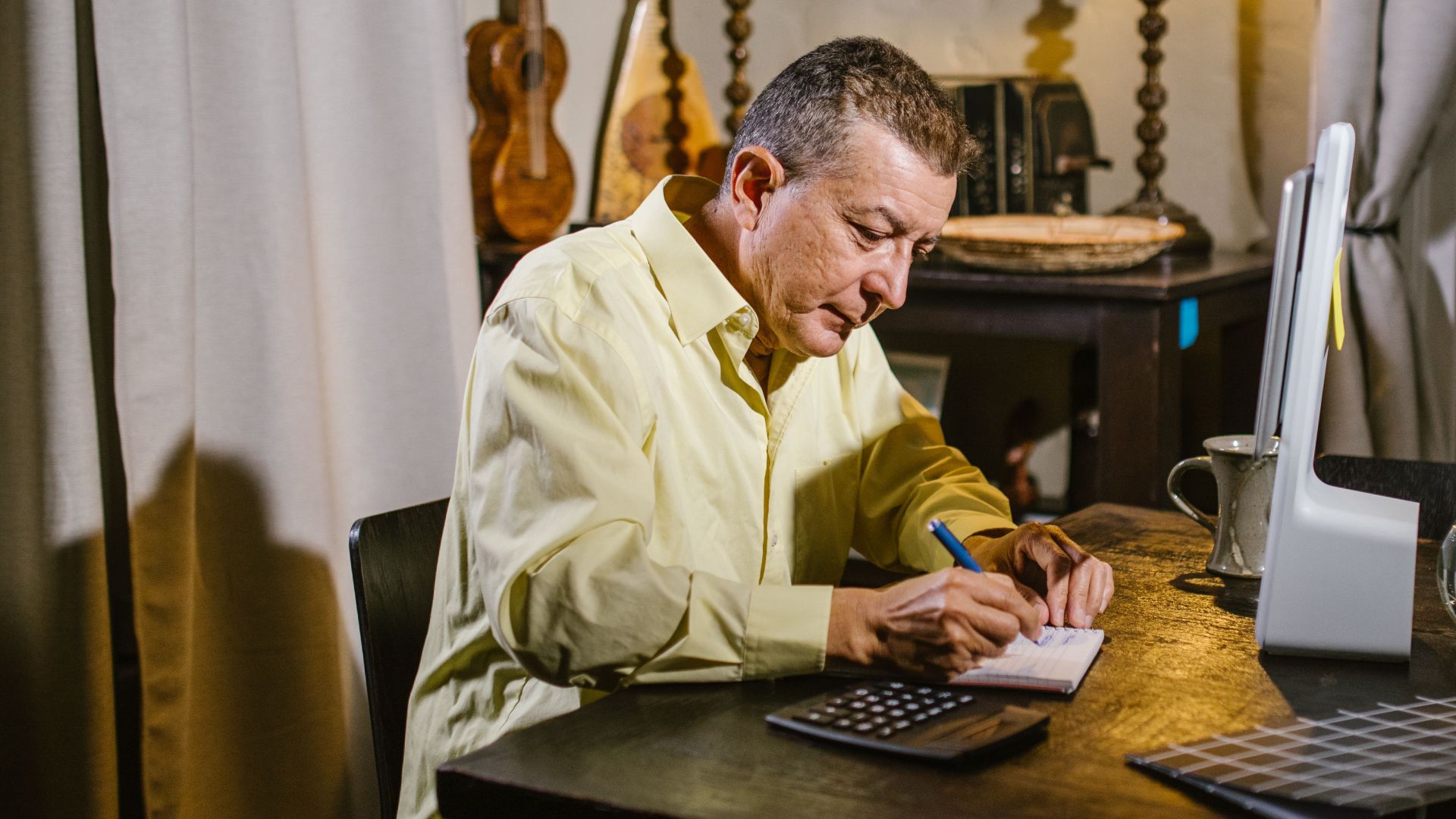 Senior man wearing yellow shirt writing notes at desk with lamp, calculator, and glass sphere nearby.