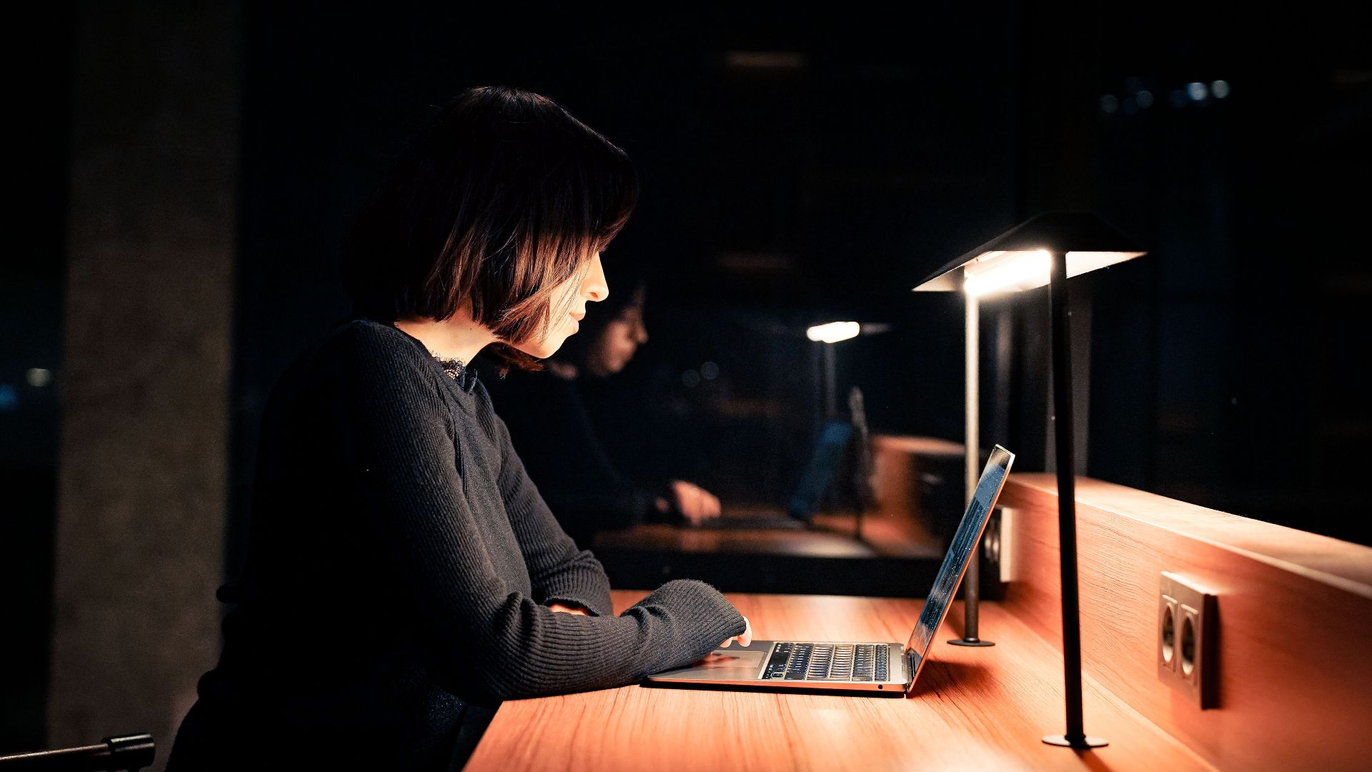 A woman focused on her laptop at a desk with dim lighting, working late at night.