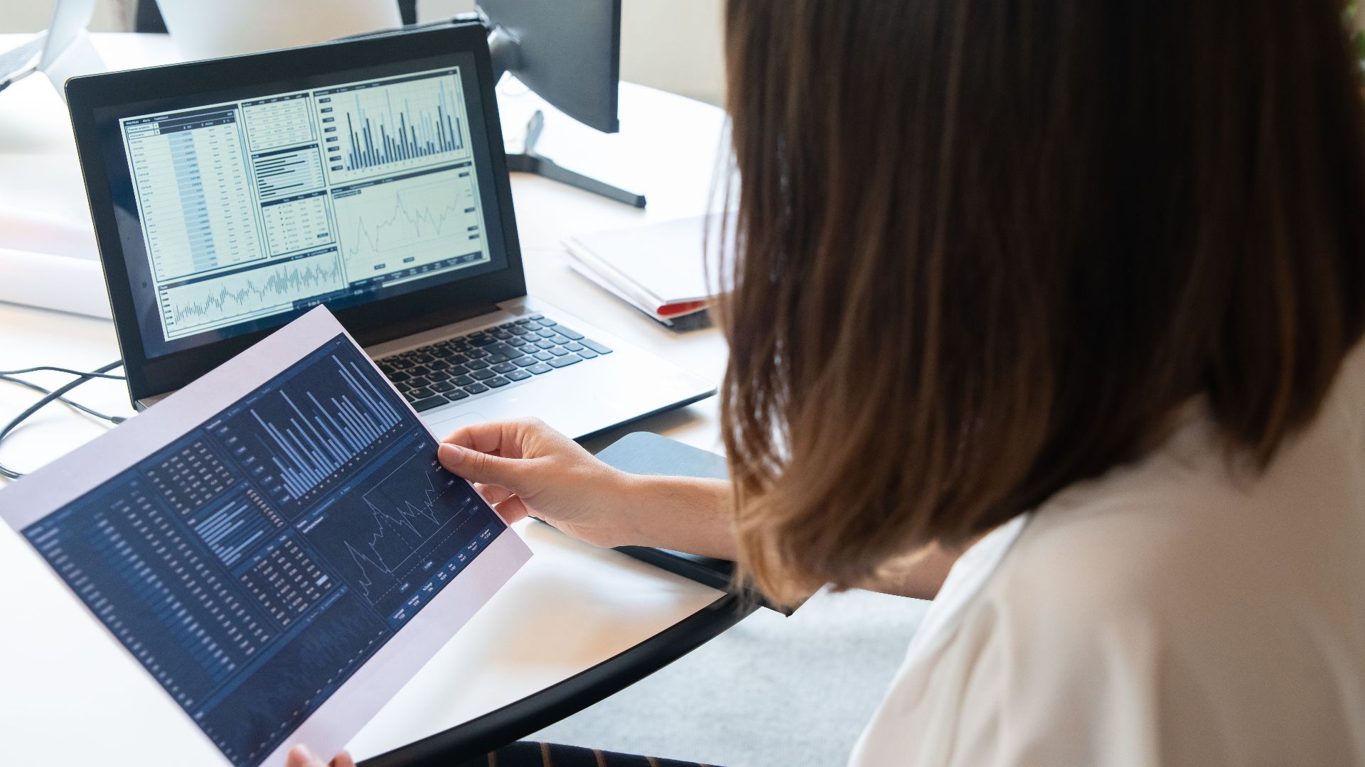 A professional woman analyzes financial charts on a laptop and paper for business insights.