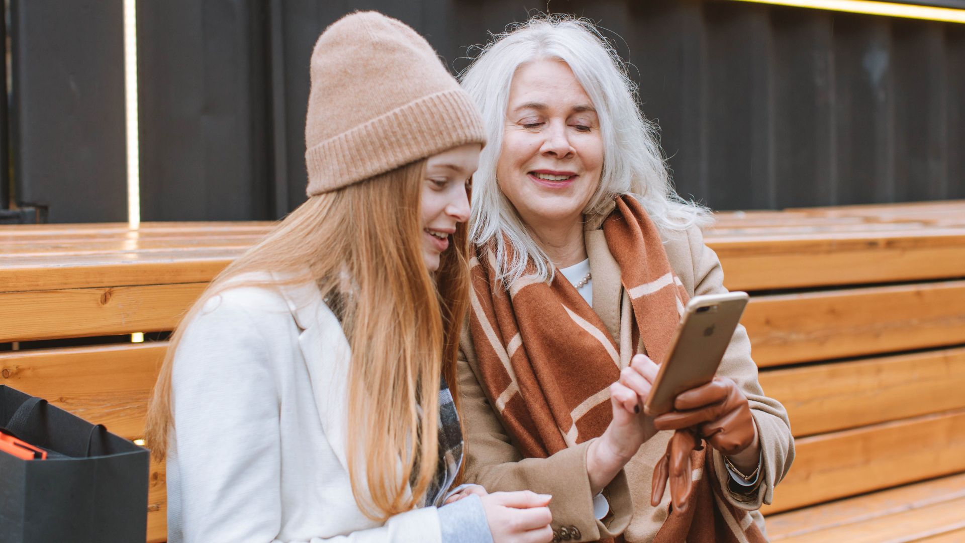 A senior woman sharing a moment with a teenager while looking at a smartphone outdoors on a wooden bench.