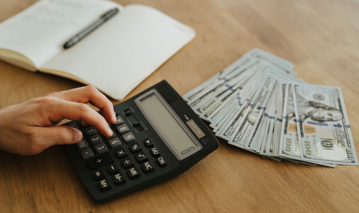 Close-up of a hand using a calculator with cash and a notebook on a wooden table