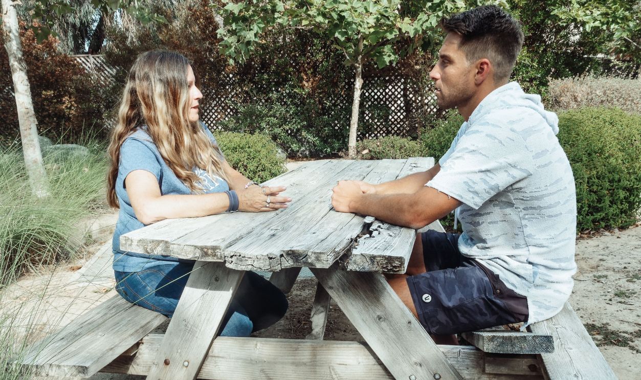 A mother and son sit talking at a rustic outdoor picnic table