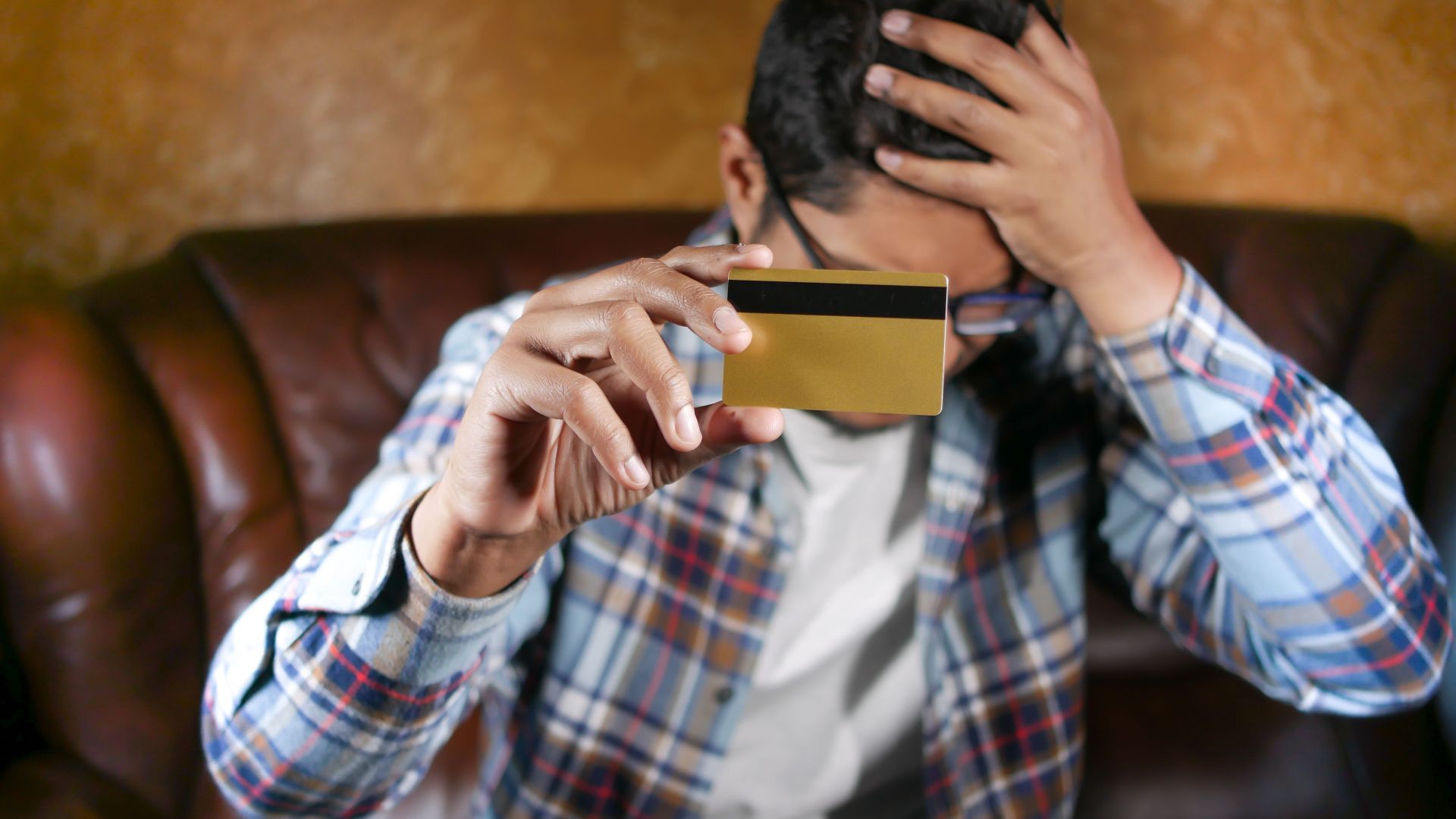 Man in checkered shirt on couch holding credit card, showing stress and worry.