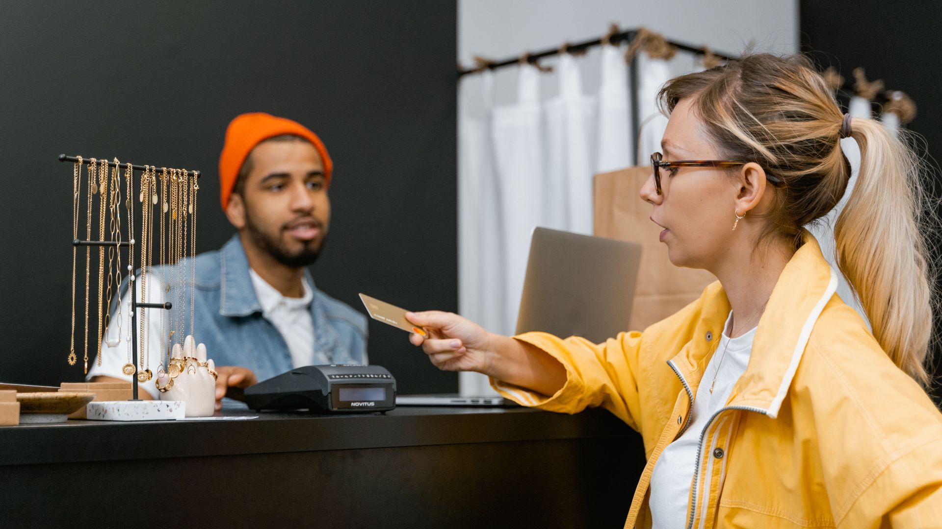 Woman in yellow jacket pays with credit card at shop counter with jewelry display.