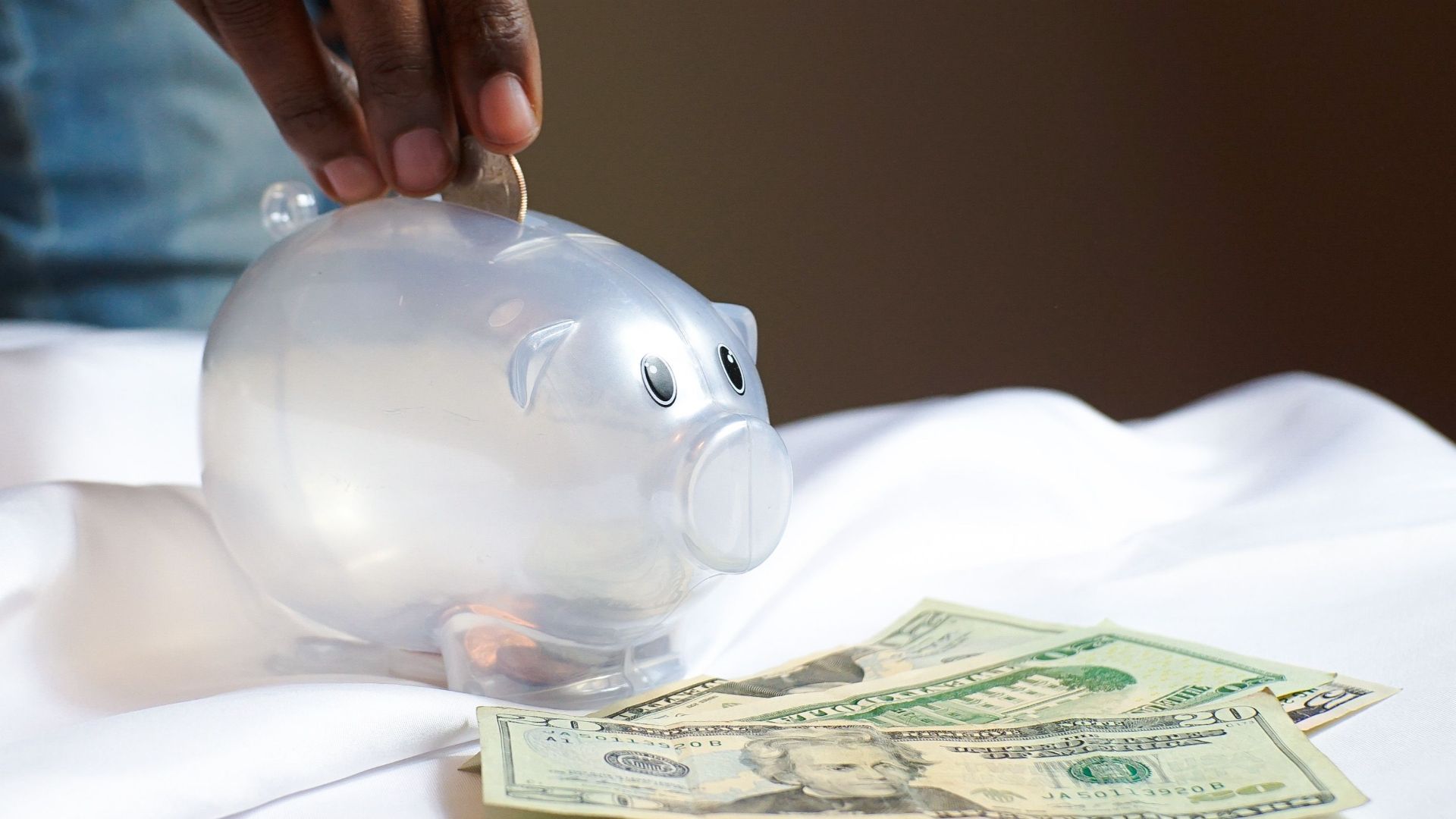 A hand placing a coin in a piggy bank with dollar bills nearby signifying savings and finance.