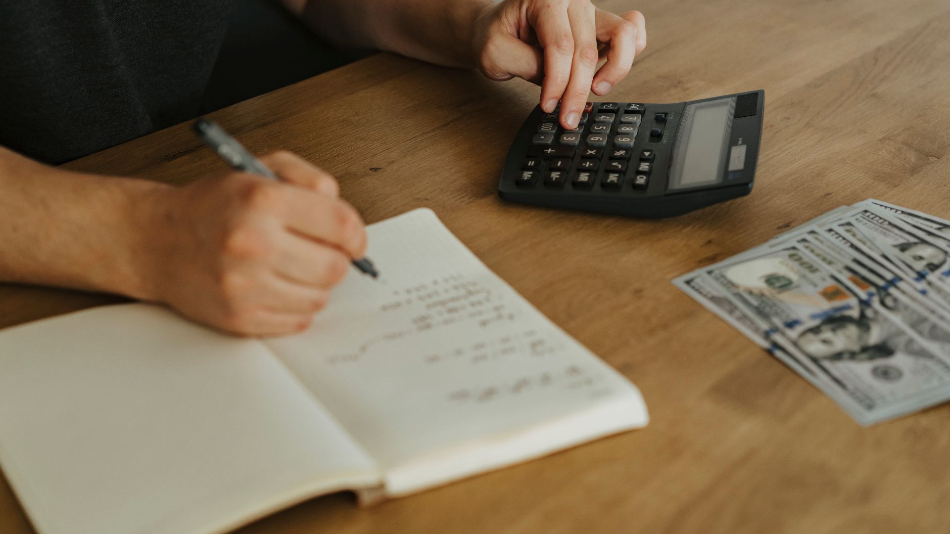 Close-up of hands using a calculator with notebook and cash on a wooden table.