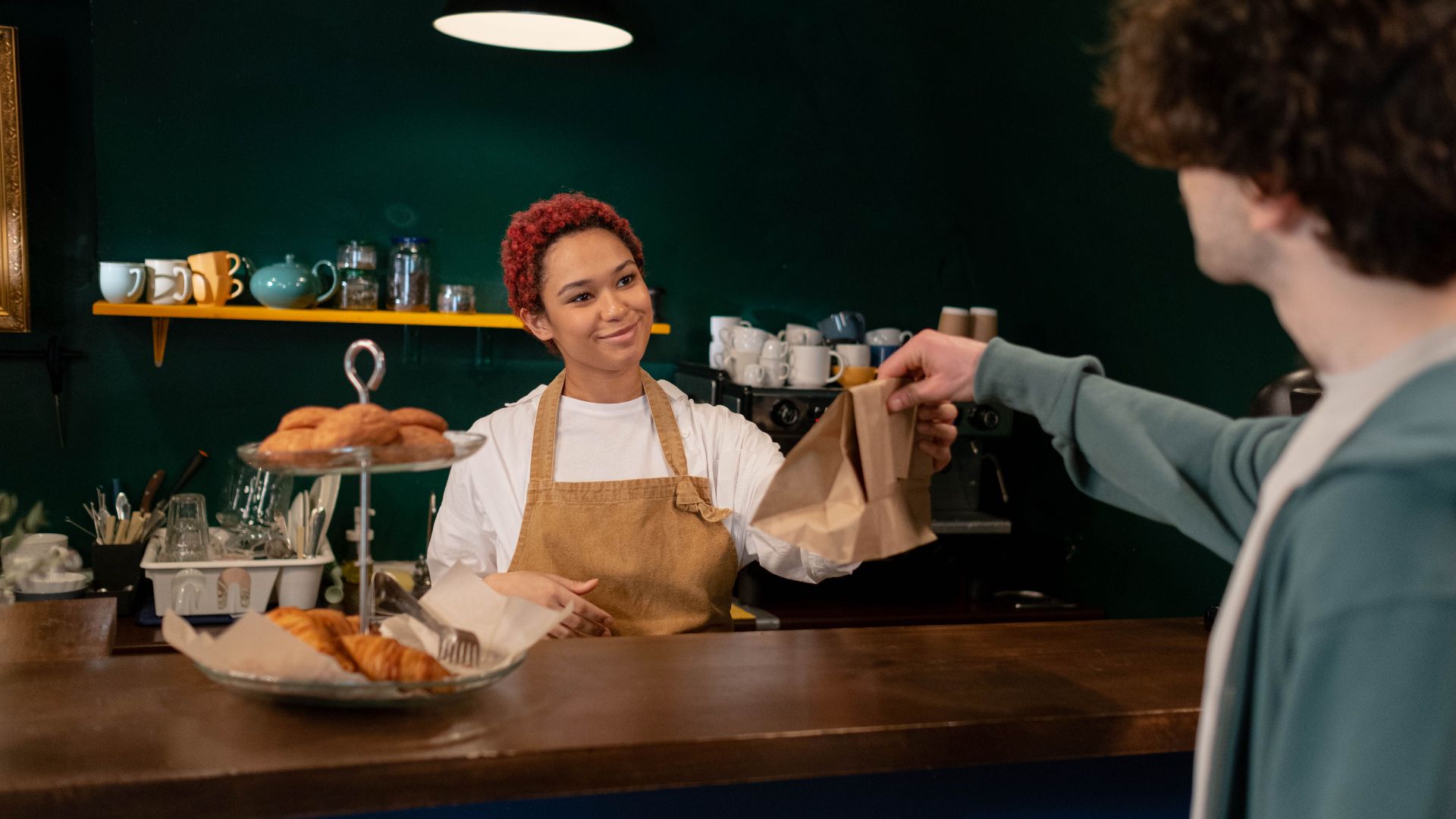 A friendly barista hands over pastries to a customer at a cozy café counter. Warm ambiance indoors.