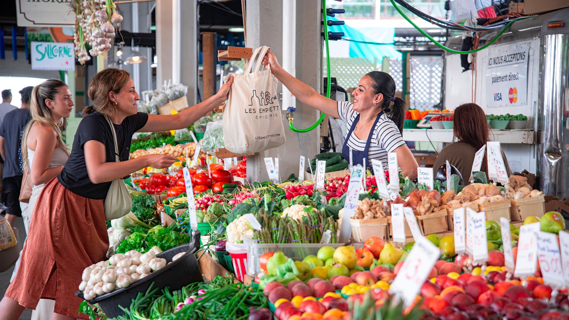 Women shopping at a colorful outdoor market stall filled with fresh fruits and vegetables. Smiles and interaction.