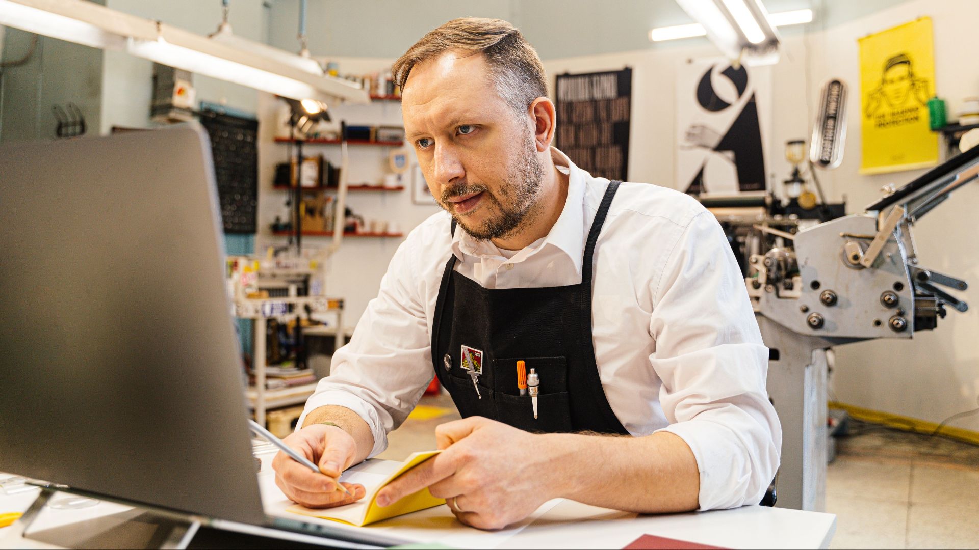 A dedicated man working at his computer within a busy print shop space, focusing intently.