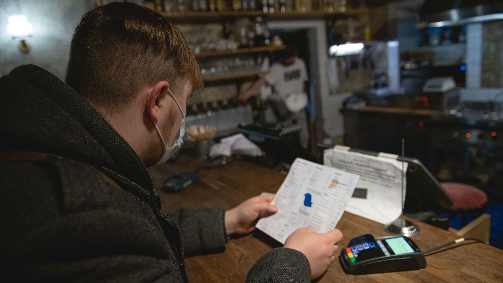 A man with a face mask reads a menu at a bar counter, creating a cozy and intimate atmosphere.