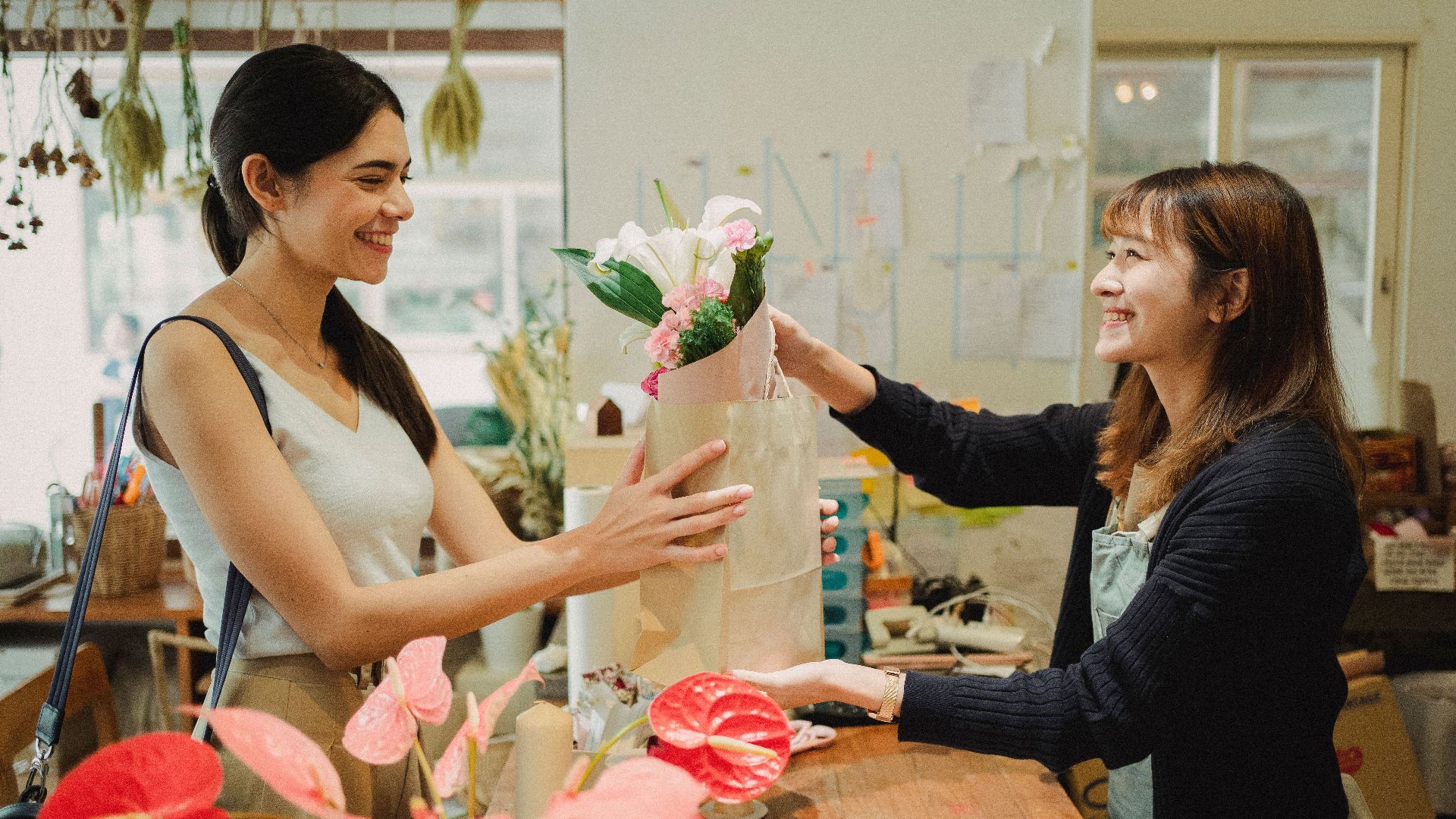 Side view of cheerful florist in apron giving paper bag with bouquets of flowers to smiling customer in floral shop in daytime