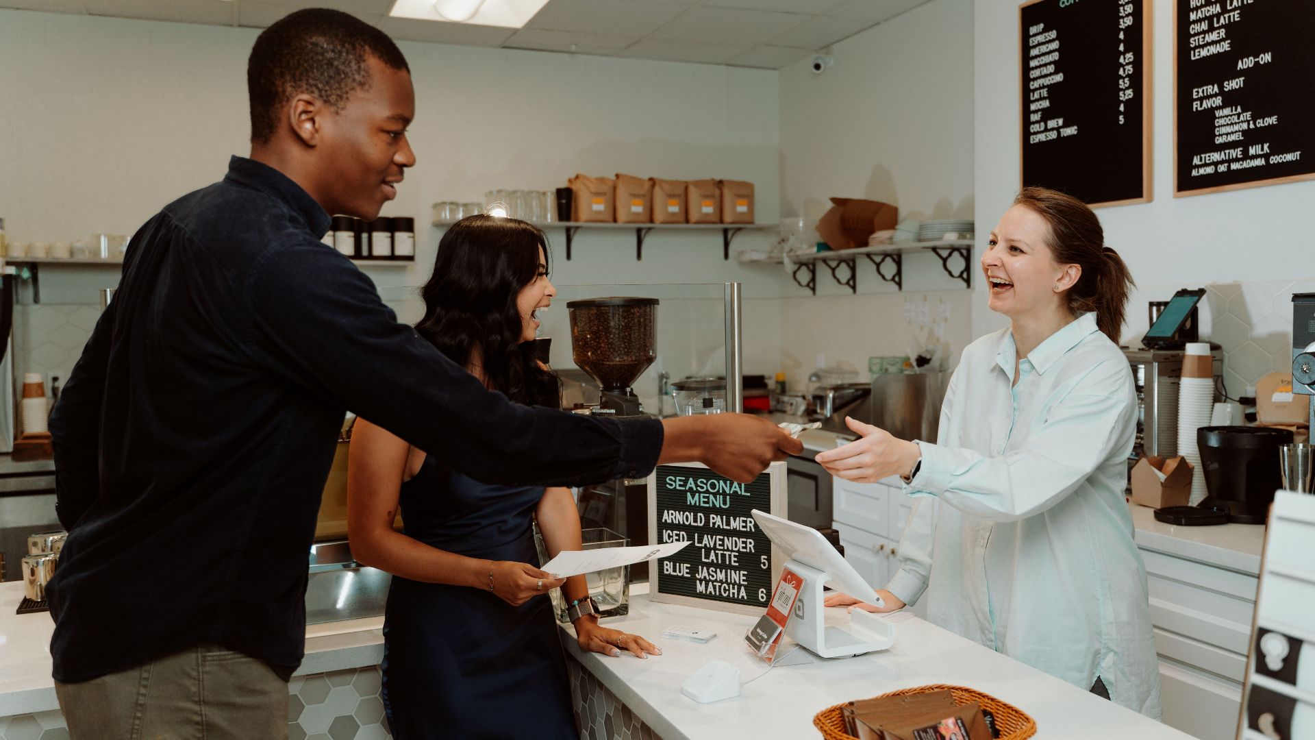 Customers and barista laughing and exchanging payment at a coffee shop counter.