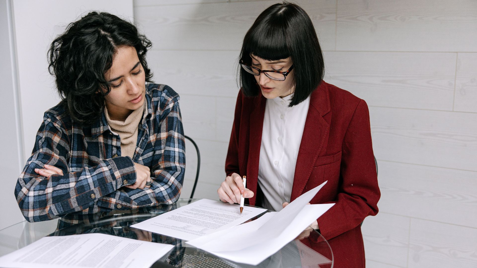 Two women collaborating at a glass table, reviewing business documents in a modern office setting.