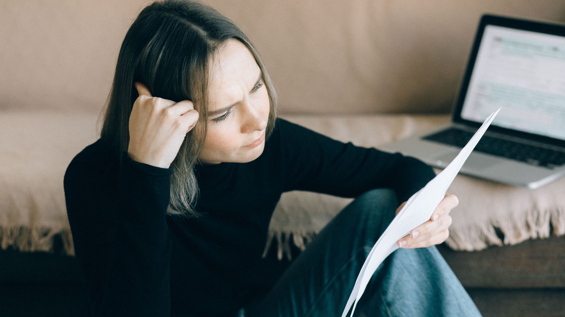 Focused woman analyzing papers with a laptop open, symbolizing thoughtful consideration.