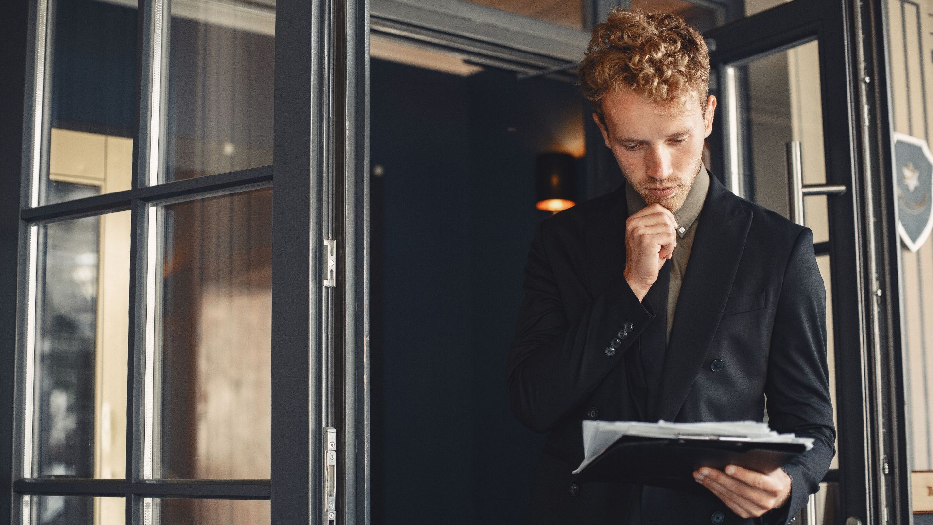 A businessman in a suit thoughtfully reviews documents while standing by a door outdoors.