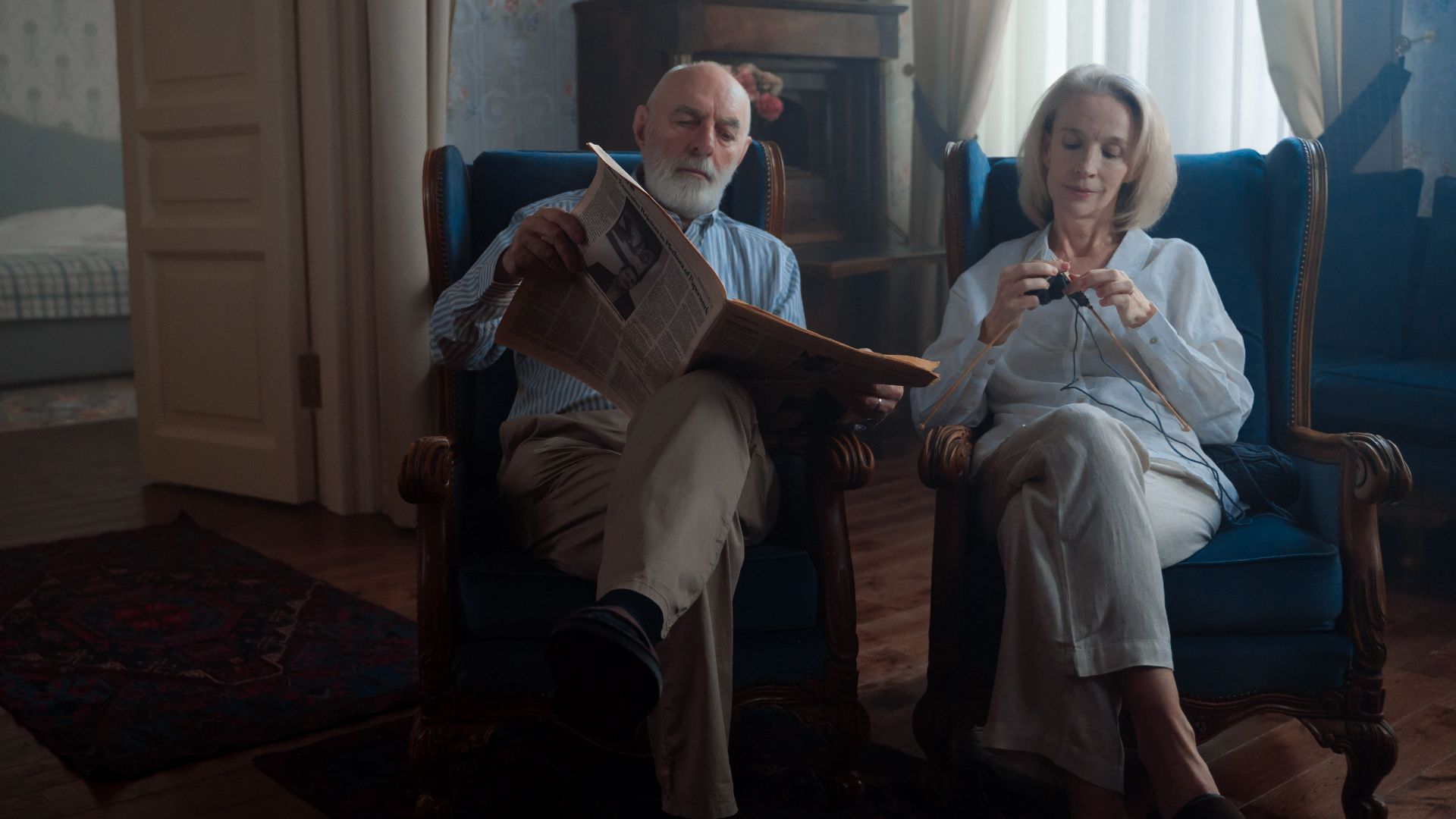 Elderly couple sitting together, one knitting, the other reading a newspaper in a cozy room.