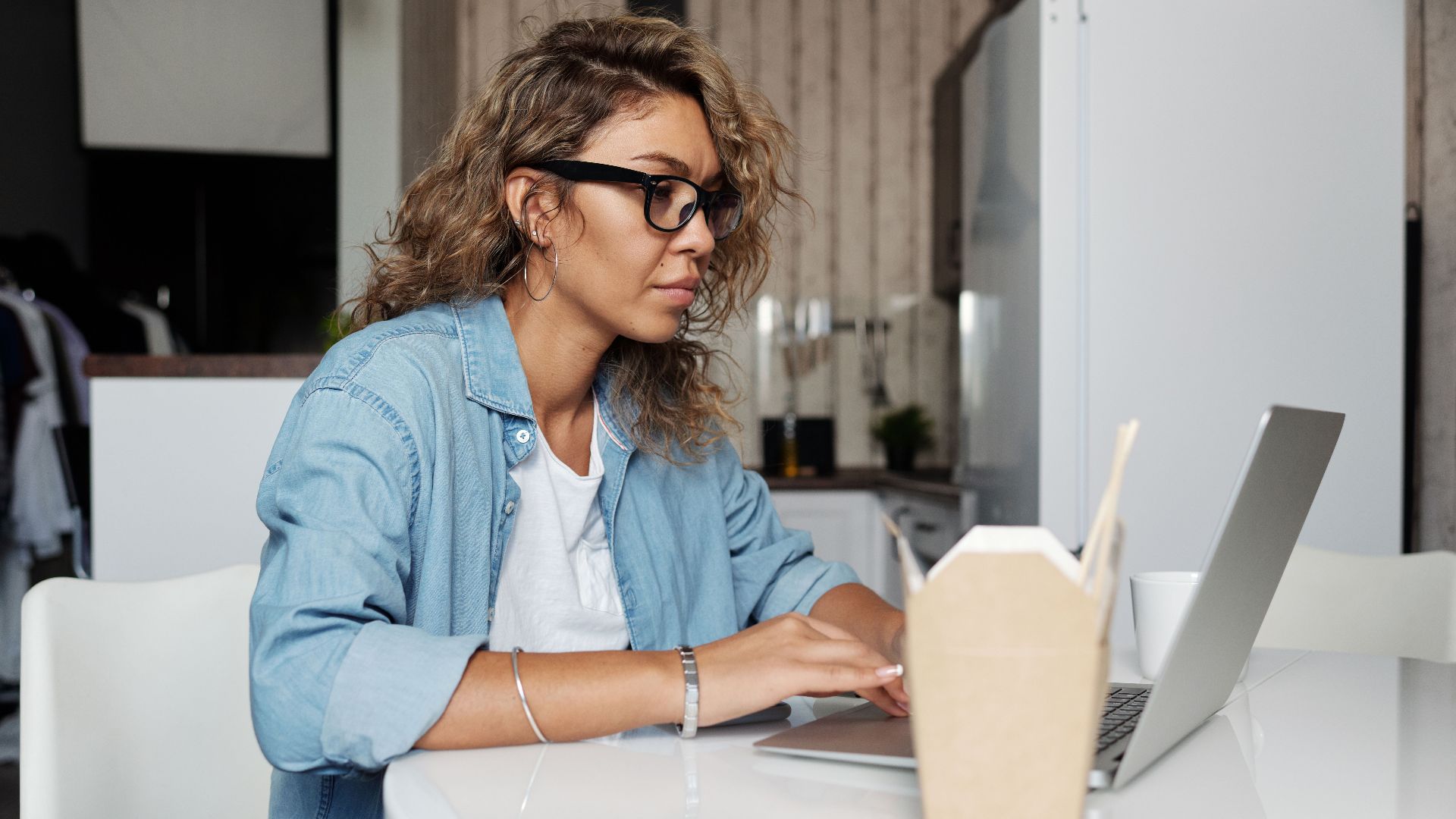 A woman in a denim jacket working remotely from home on her laptop, enjoying a casual workspace.