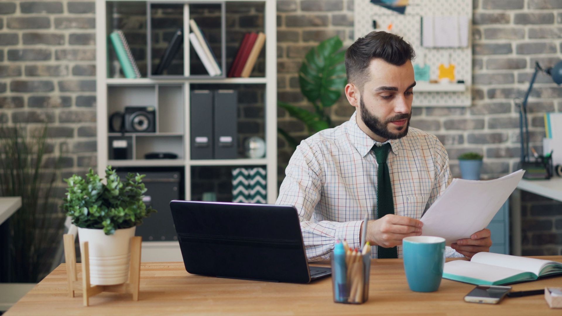 Young man reading papers at desk with laptop and office decor, focused on work.