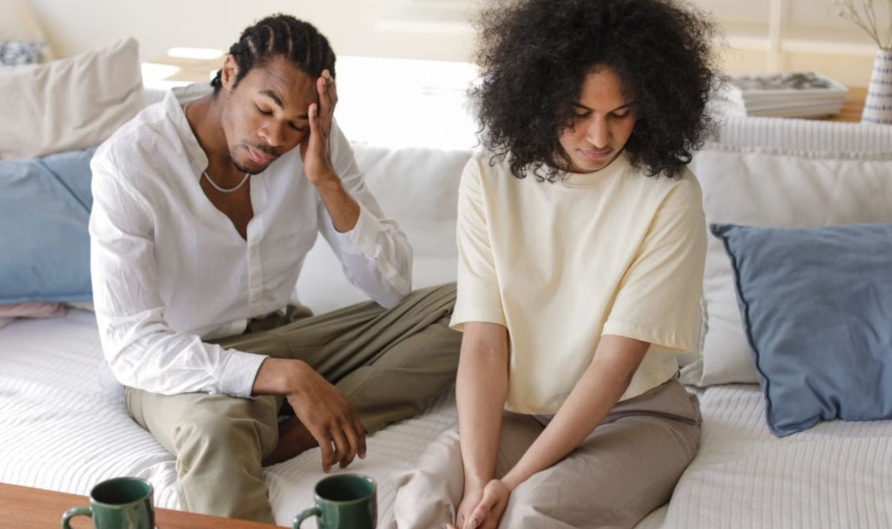 A contemplative black couple sitting on a sofa indoors in a modern setting, reflecting emotions