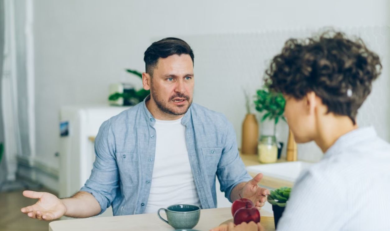 Two adults engage in a conversation at a kitchen table with coffee cups