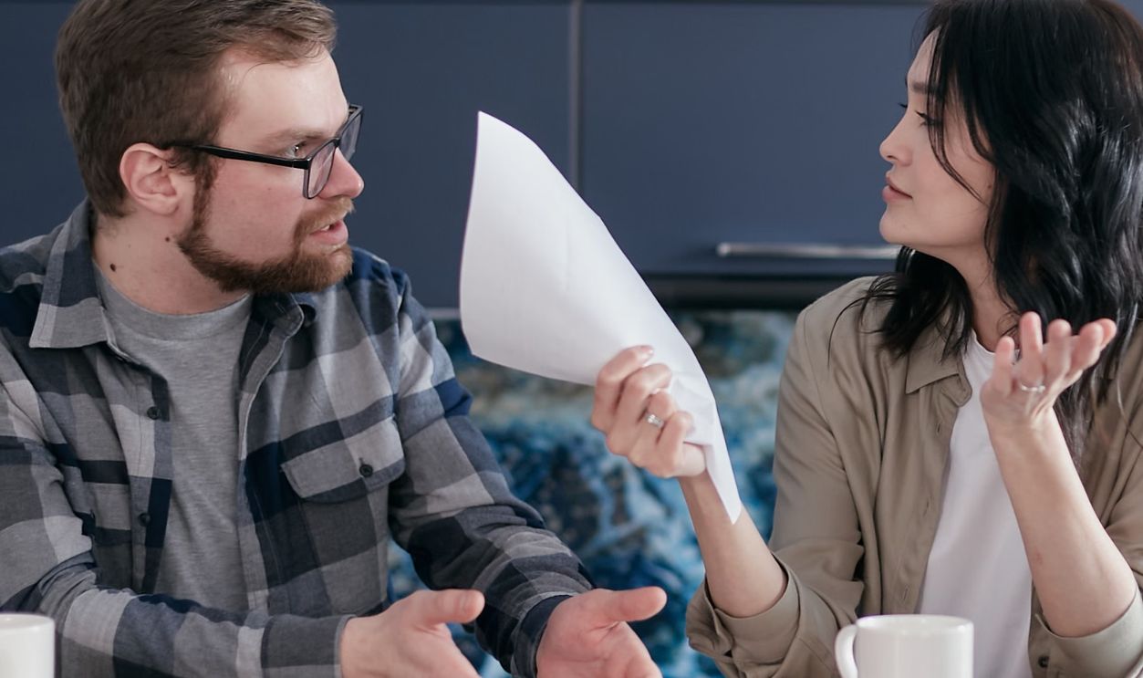 A couple sits at a table, discussing finances with papers and a laptop, looking concerned