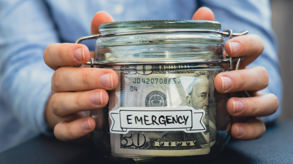 Female hands Saving Money In Glass Jar filled with Dollars banknotes. EMERGENCY transcription in front of jar. Managing personal finances extra income for future insecurity