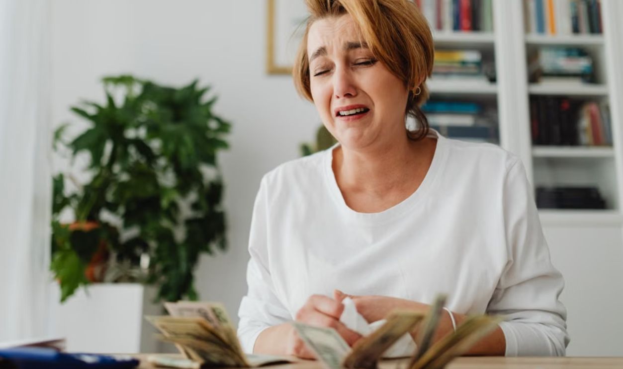 A distressed woman counts cash at a desk with a pained expression, highlighting financial strain