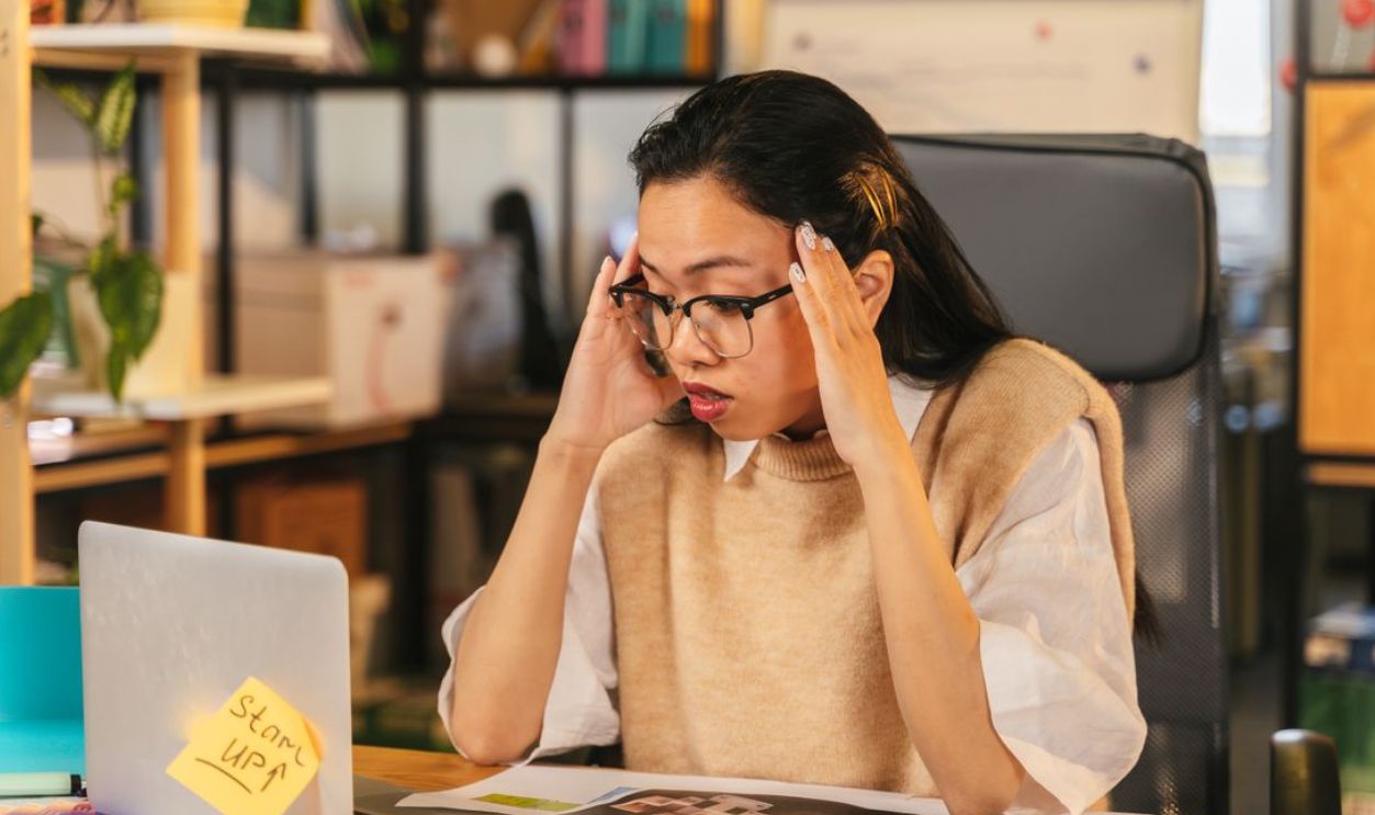 A woman with glasses shows stress while working at a laptop in an office environment