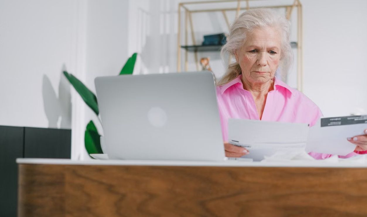 Elderly woman focused on paperwork at home office with laptop