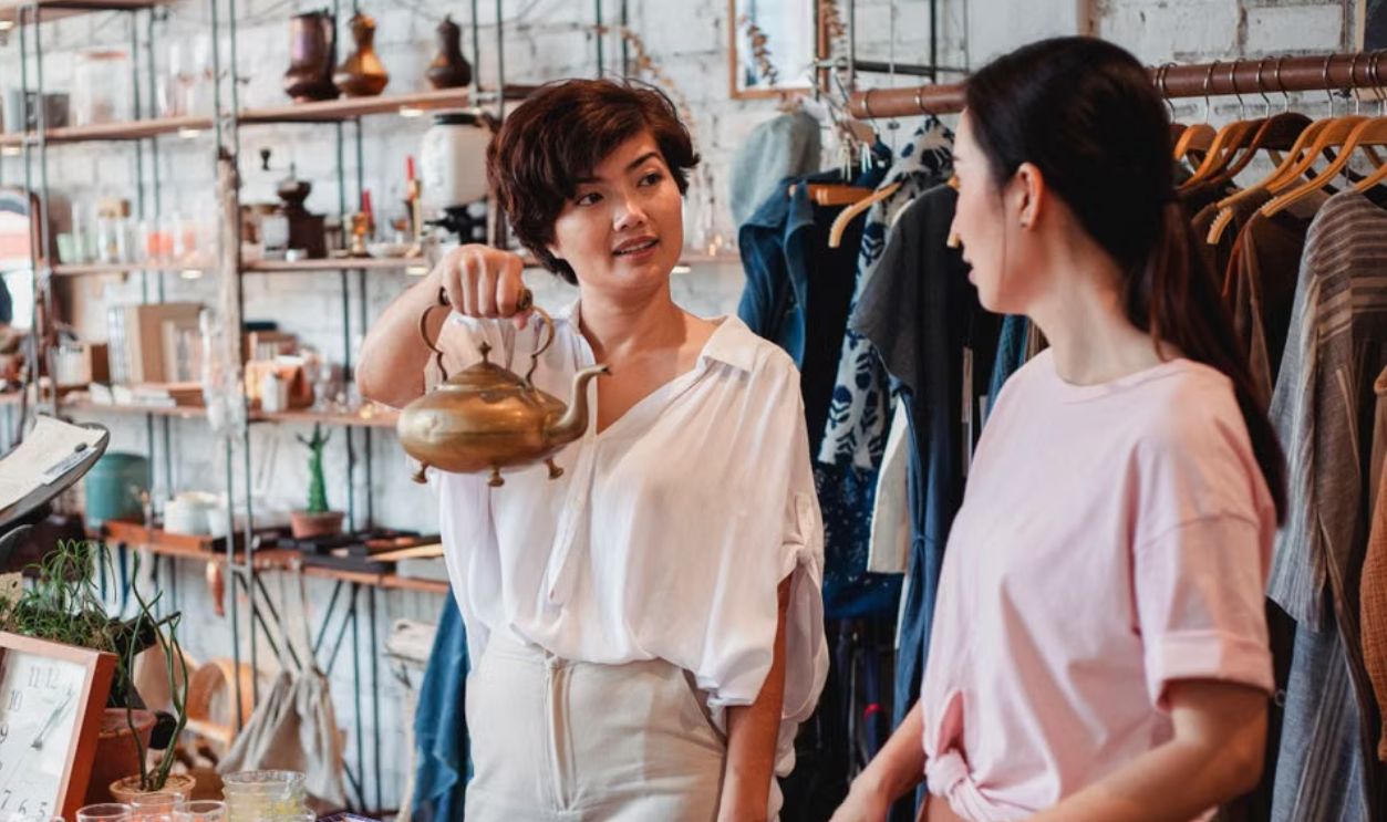 Two women shopping at a vintage store discussing a brass kettle