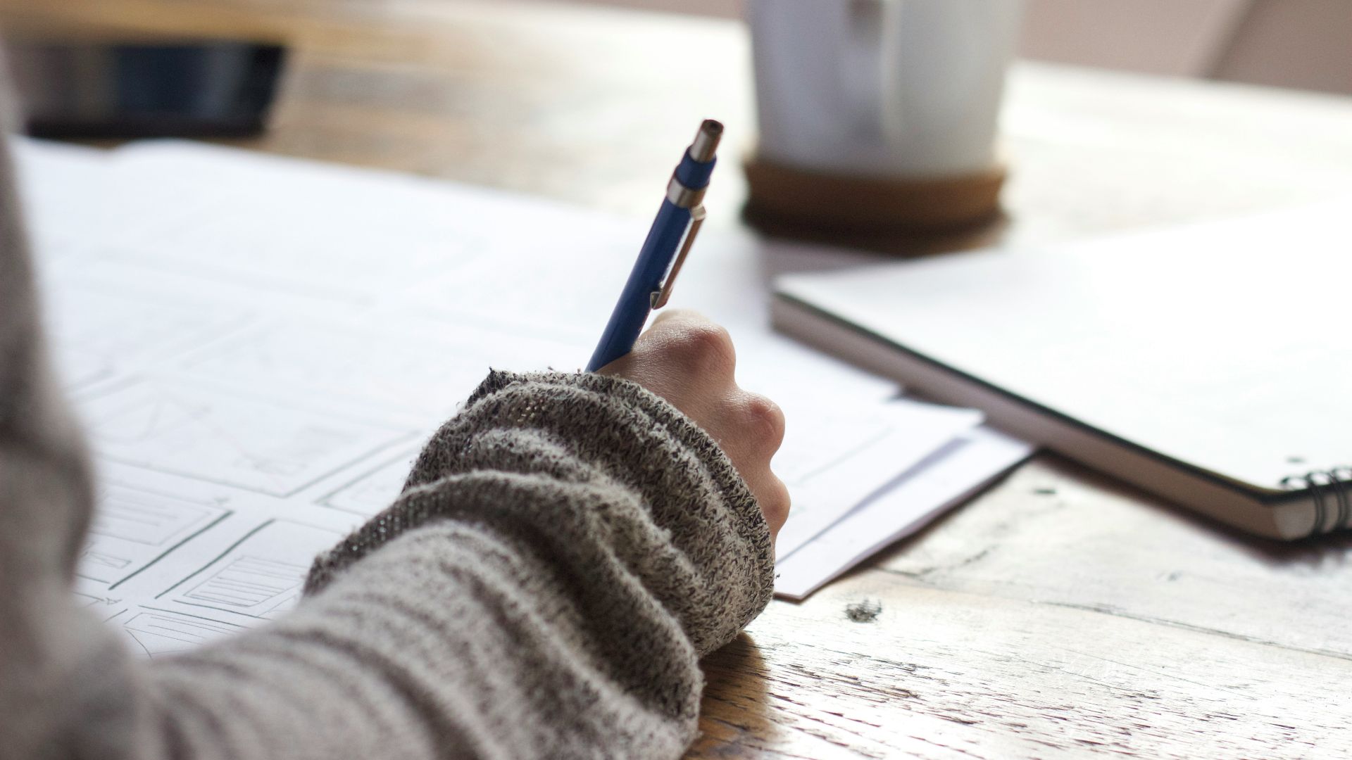 person writing on brown wooden table near white ceramic mug