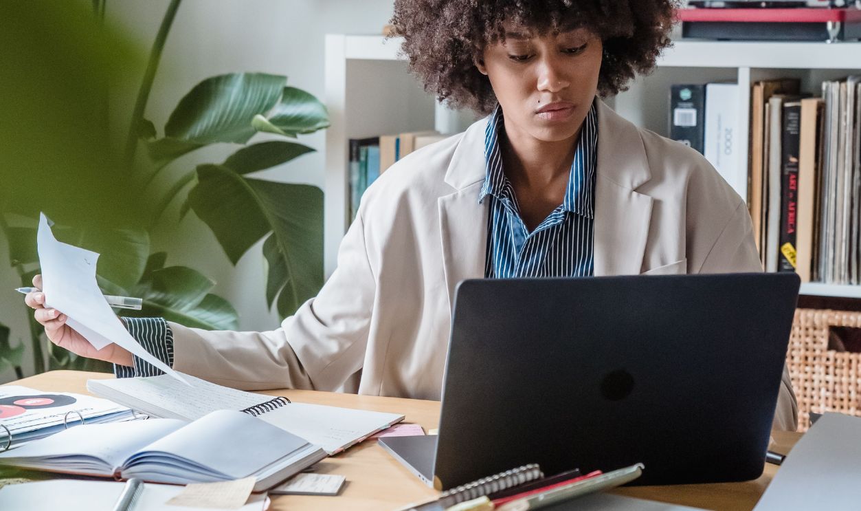 Woman working at a home office desk with a laptop, plants, and paperwork around