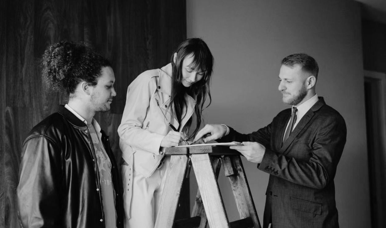 Three individuals signing a contract on a ladder inside a new house, symbolizing a real estate deal.