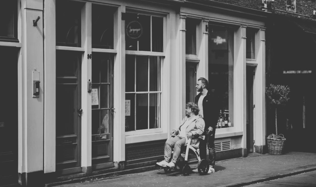 A man helps a woman in a wheelchair along a picturesque street in Amsterdam, Netherlands.