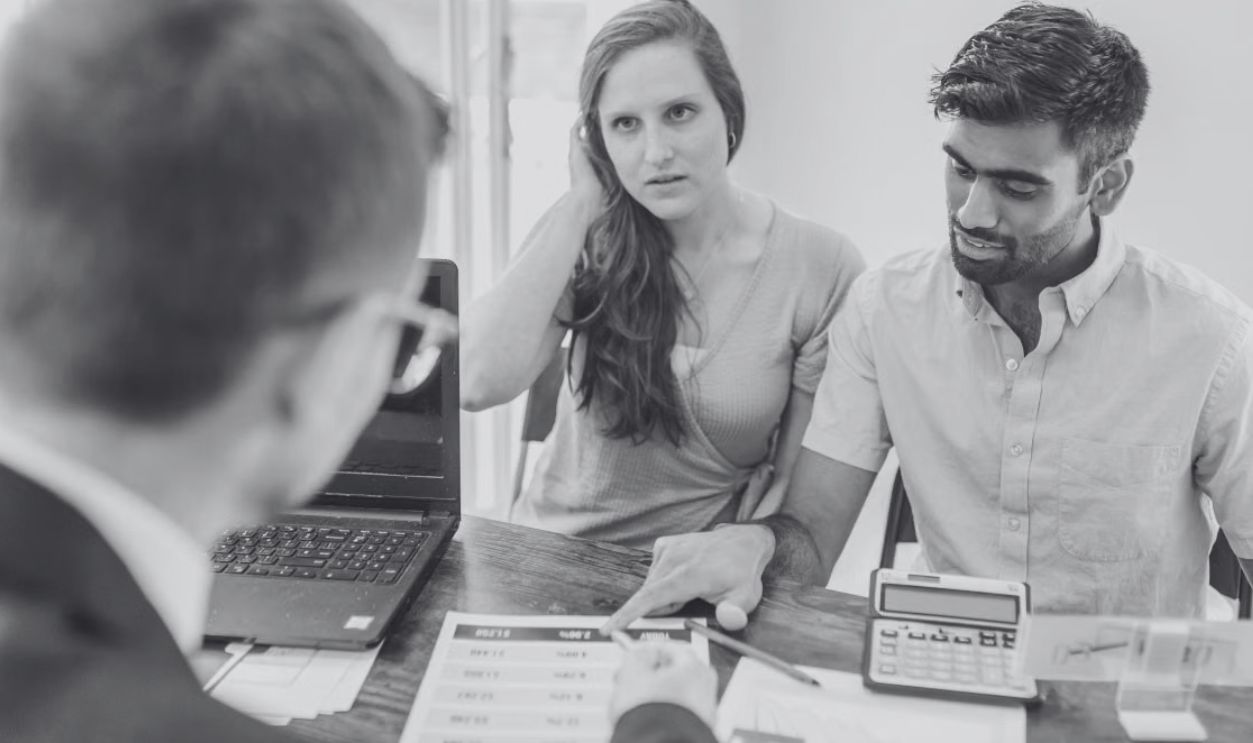 A young couple consults with a realtor about buying a home, indoors in a modern office setting.