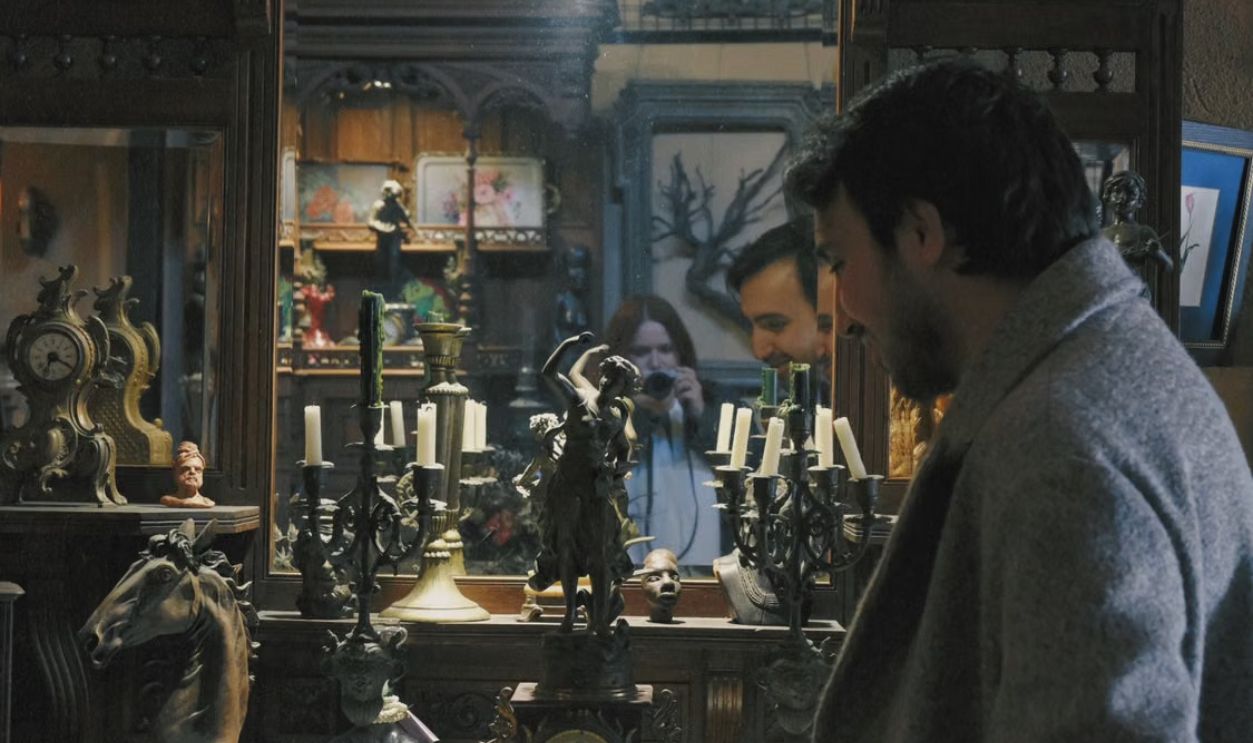 A man captured in a mirror reflection surrounded by vintage candelabras and statues in an antique shop