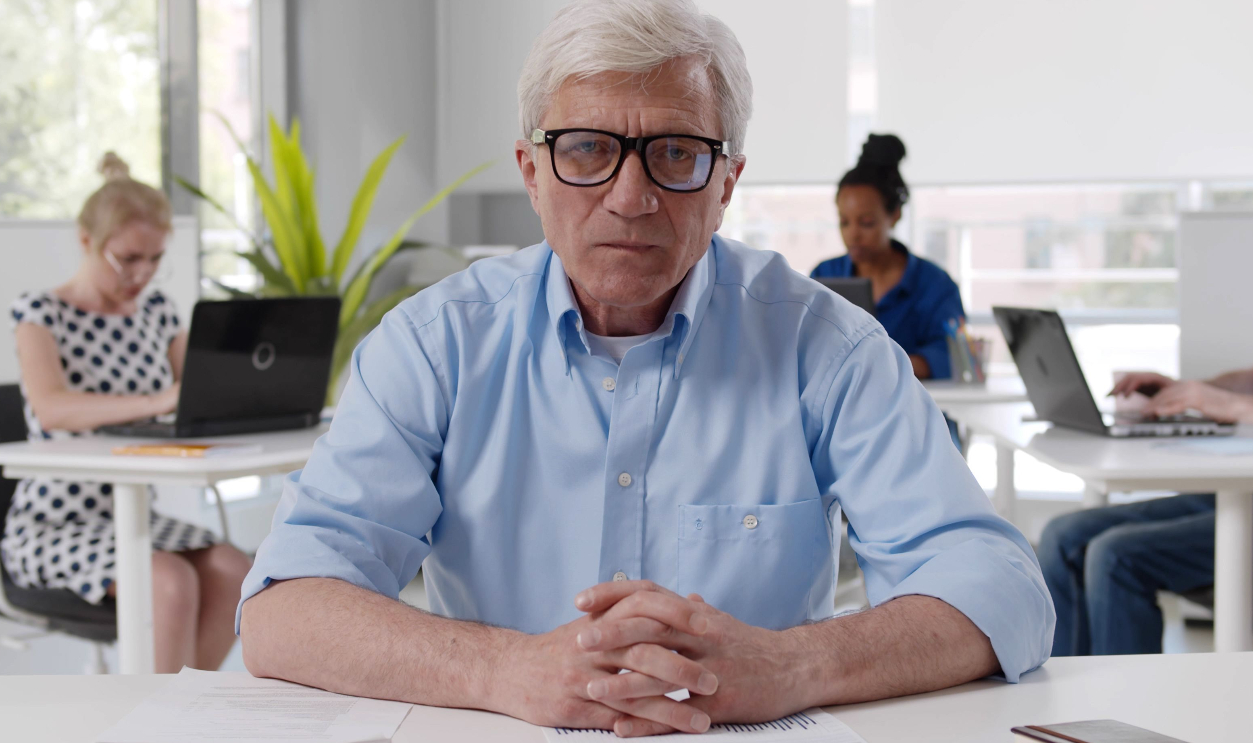 Senior manager sitting at desk in office with diverse colleagues working on background