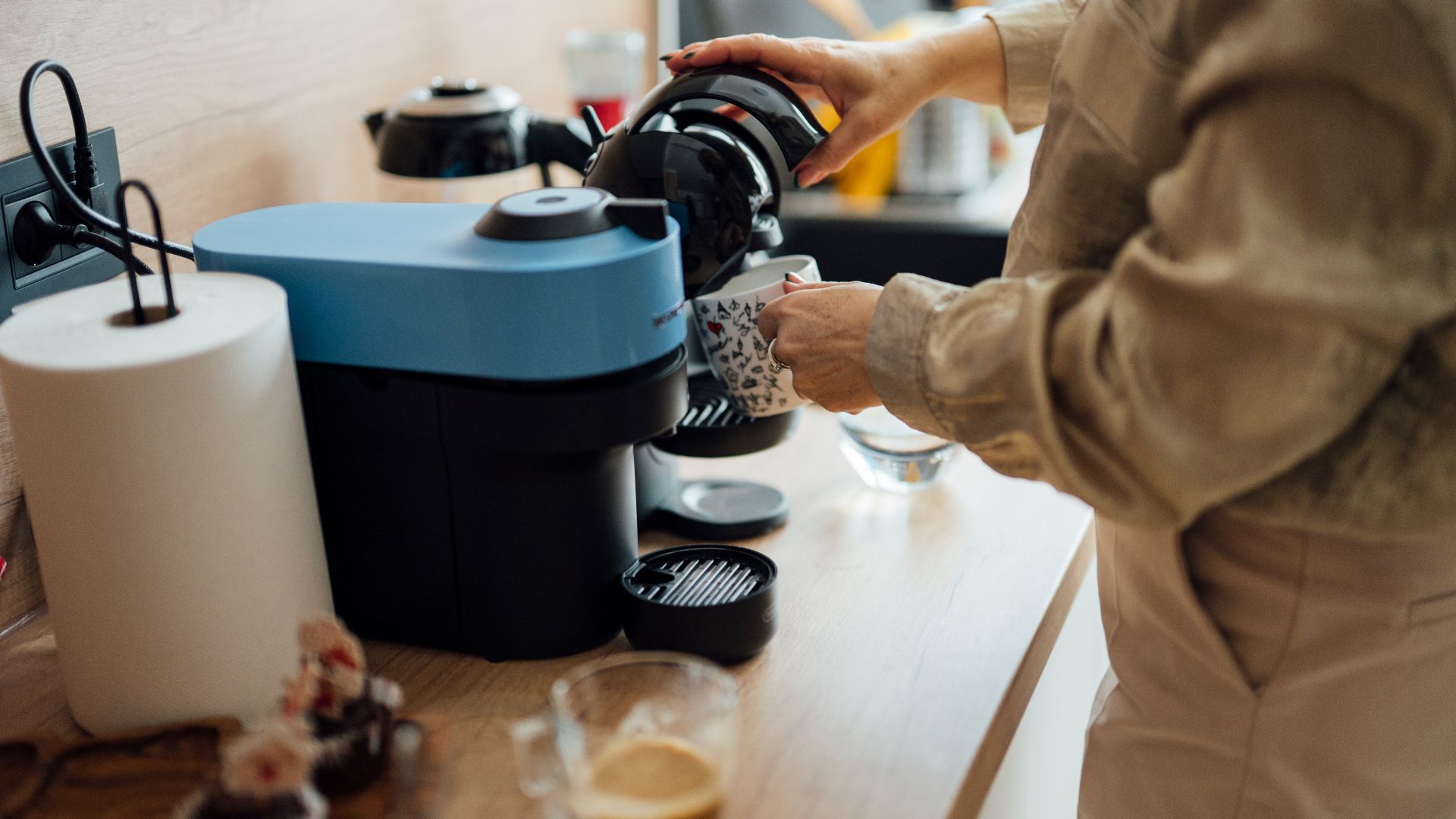 A person pours coffee from a machine into a cup in a bright kitchen. The countertop has a coffee maker, glasses, and sweet treats nearby. It looks like morning time.