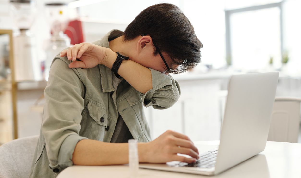 A man sneezes into his elbow while using a laptop, depicting common cold symptoms