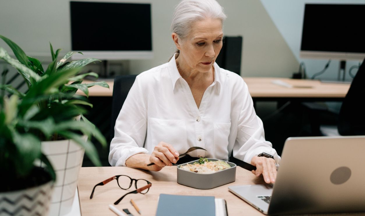Elderly woman multitasking with lunch and work on laptop in an office
