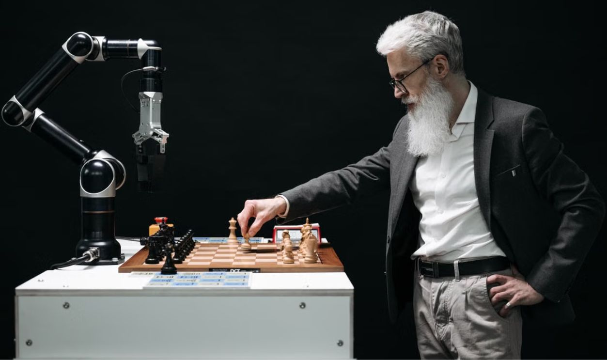 A bearded man engages in a chess game with a robotic arm, highlighting technology and strategy.