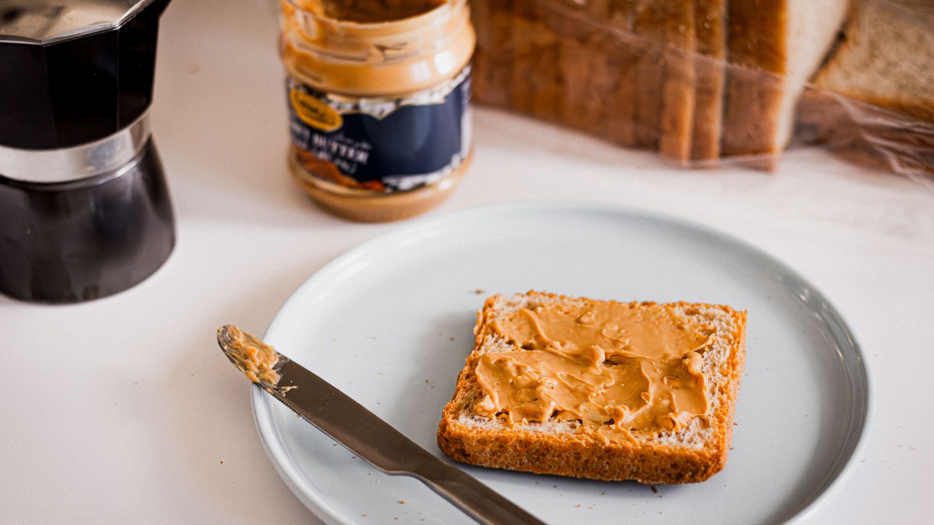 A slice of bread with peanut butter and a moka pot, perfect for breakfast.