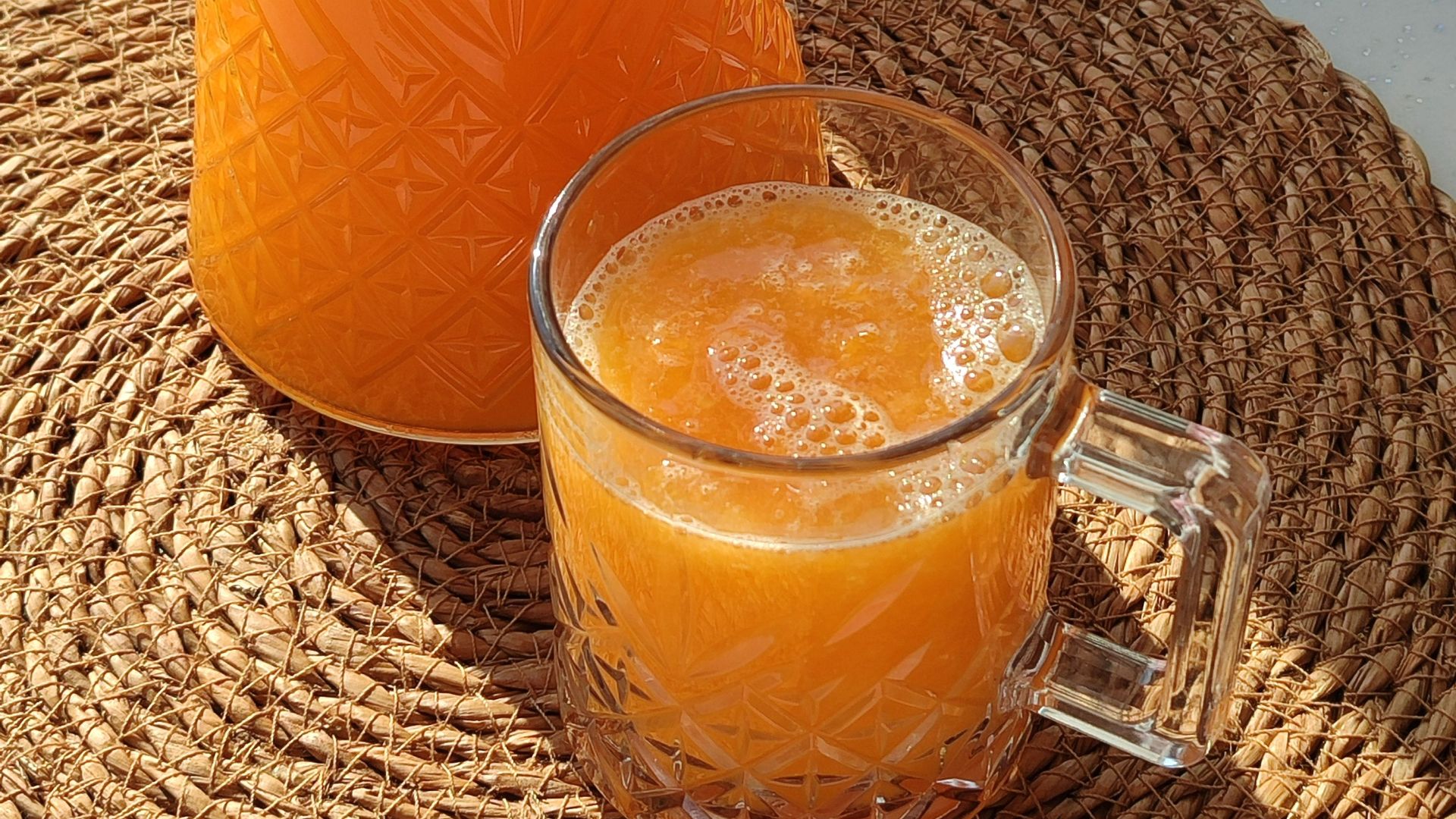 Crystal pitcher and glass of fresh orange juice basking in sunlight on woven mat.