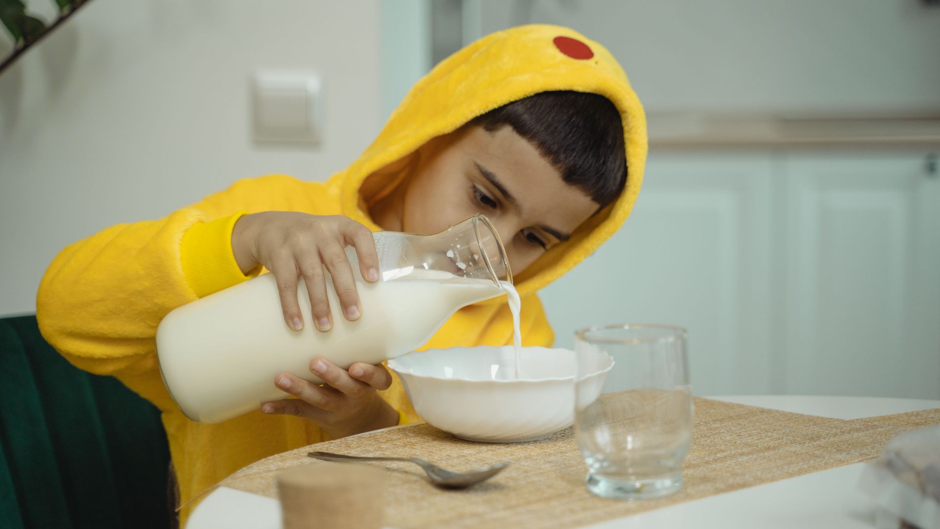 A child in a yellow hoodie carefully pouring milk into a bowl at the table.