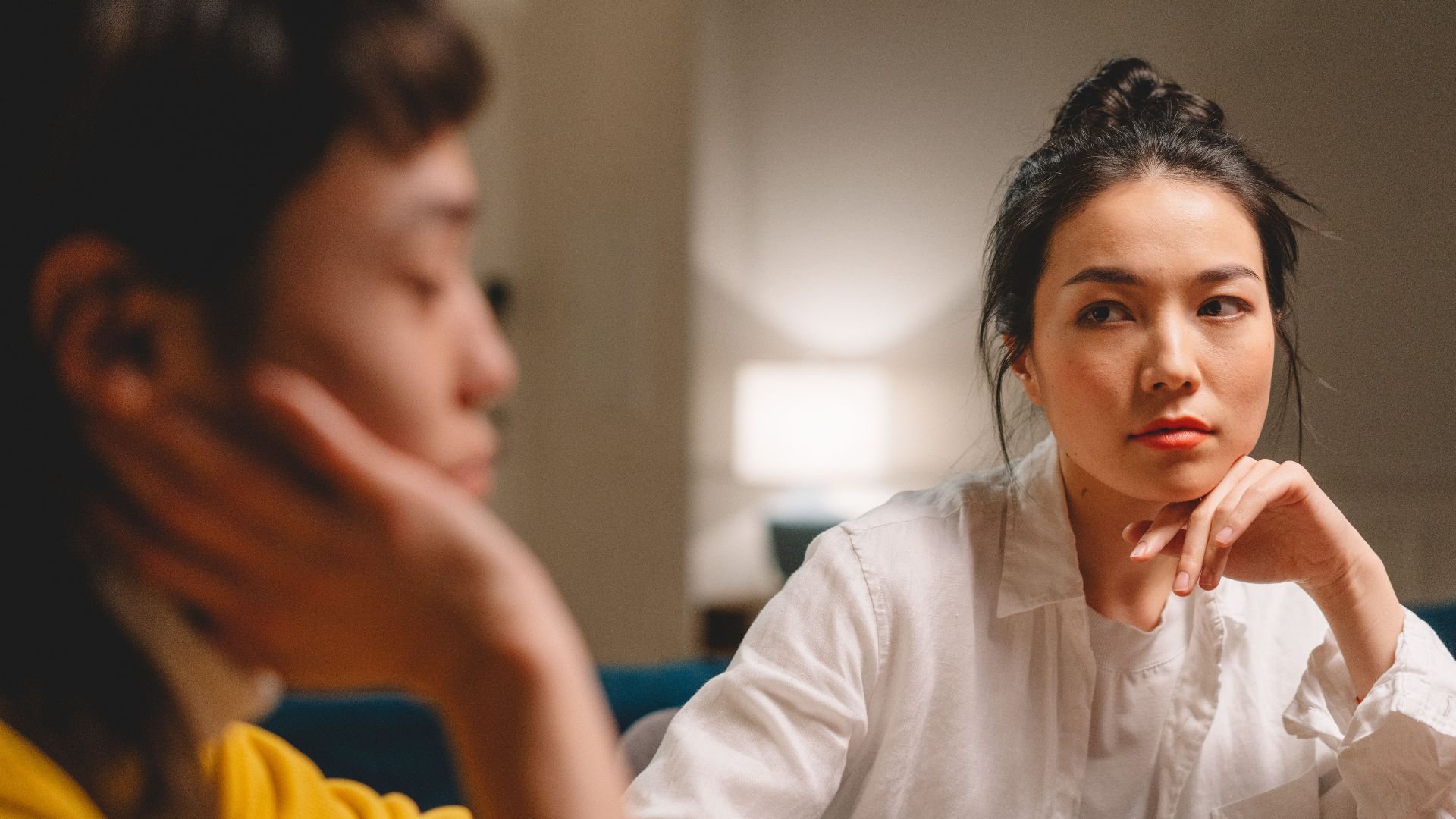 Serious concentrated young Asian female friends with dark hair in casual clothes sitting at table with hands at chins and discussing problems at home