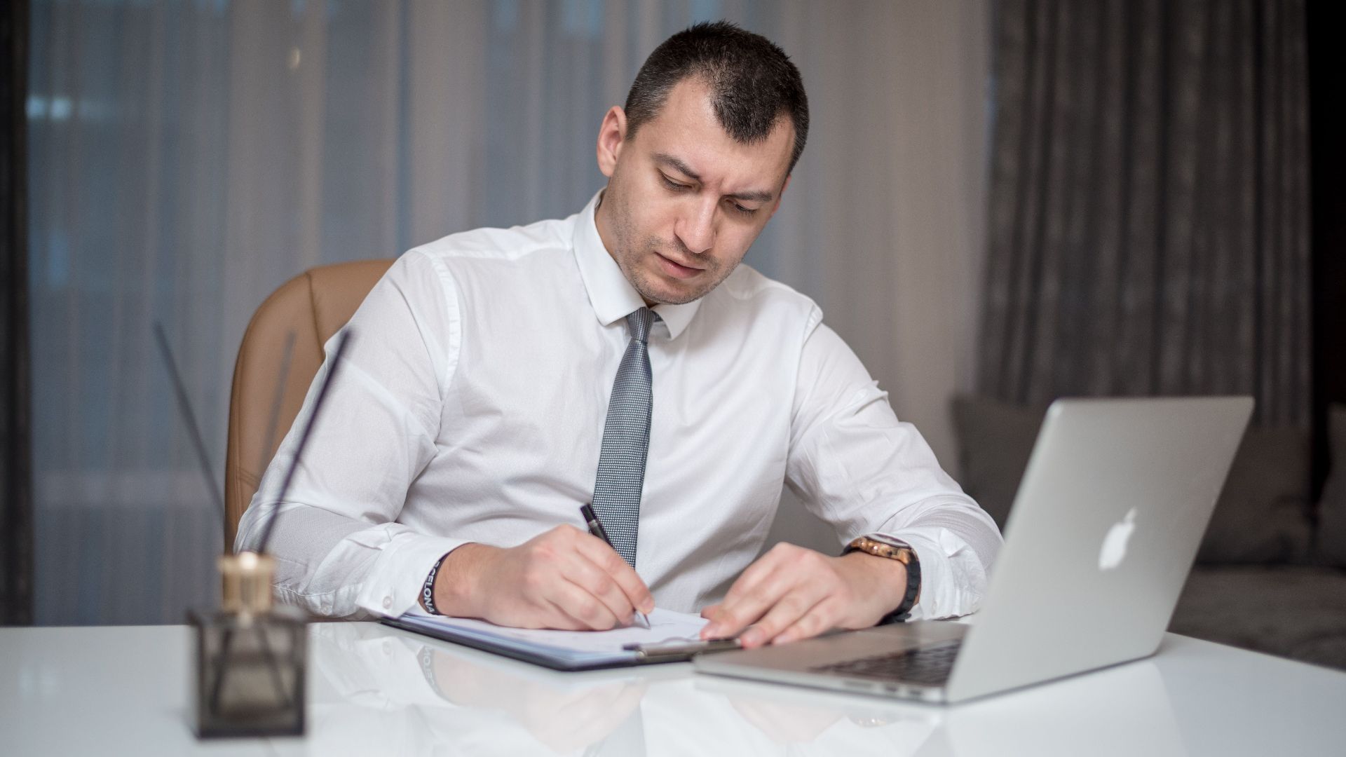 Businessman in suit writing notes at desk with laptop, reflecting modern office life.
