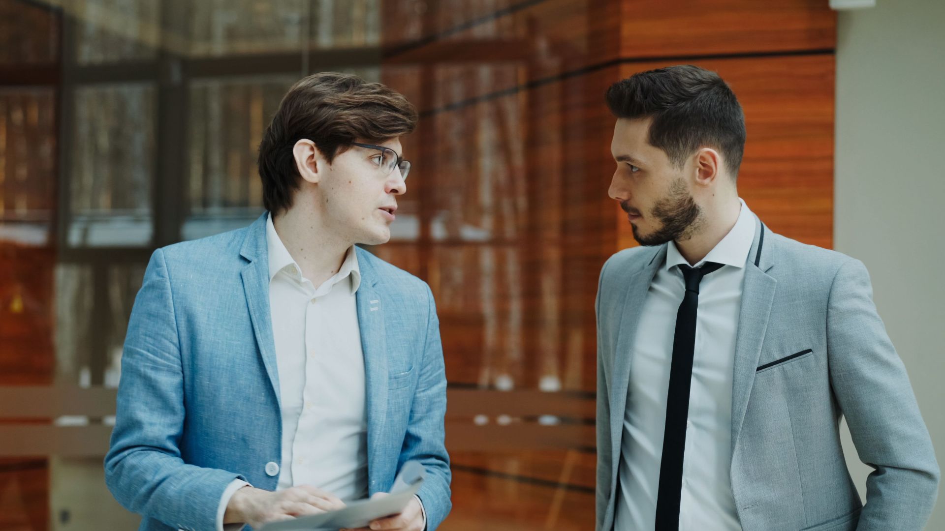 Two businessmen in suits having a conversation indoors, modern office setting.